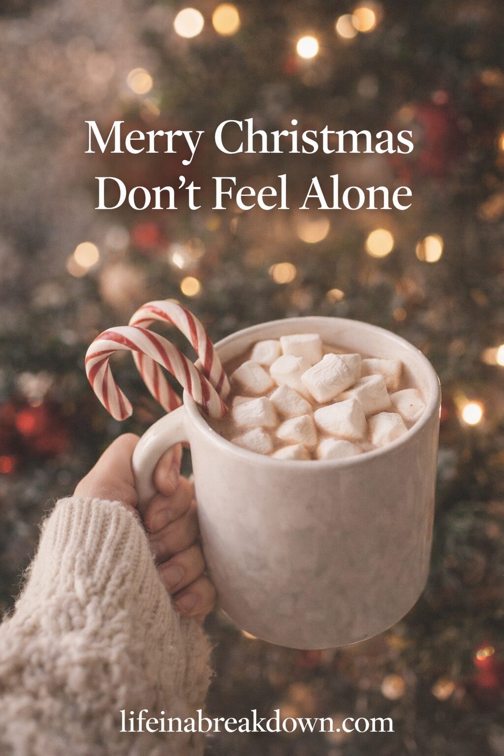 A hand holds a mug of hot chocolate topped with marshmallows and two candy canes in front of a blurred, decorated Christmas tree. Text reads: “Merry Christmas Don’t Feel Alone.”.
