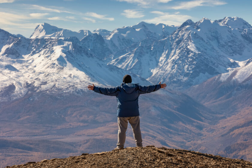 View of a tourist spreading his arms and facing the mountains. White snowy mountain ridge and beautiful blue cloudy sky as a background and slightly out of focus. Altai mountains, Siberia, Russia.