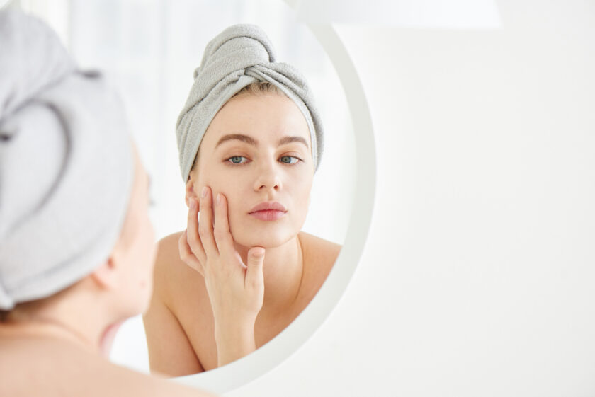 Portrait of young girl with towel on head in white bathroom looks and touches her face in the mirror and enjoys youth and hydration. Natural beauty, home care for problem skin