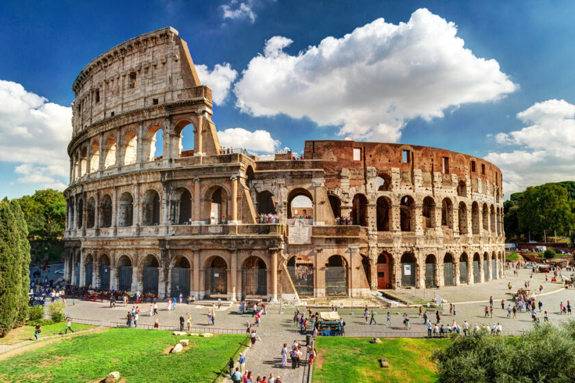 The image shows the Colosseum in Rome, Italy, under a clear blue sky with scattered clouds. Tourists are walking around the ancient amphitheater, highlighting its grand architecture and historical significance. Green trees are visible in the background.