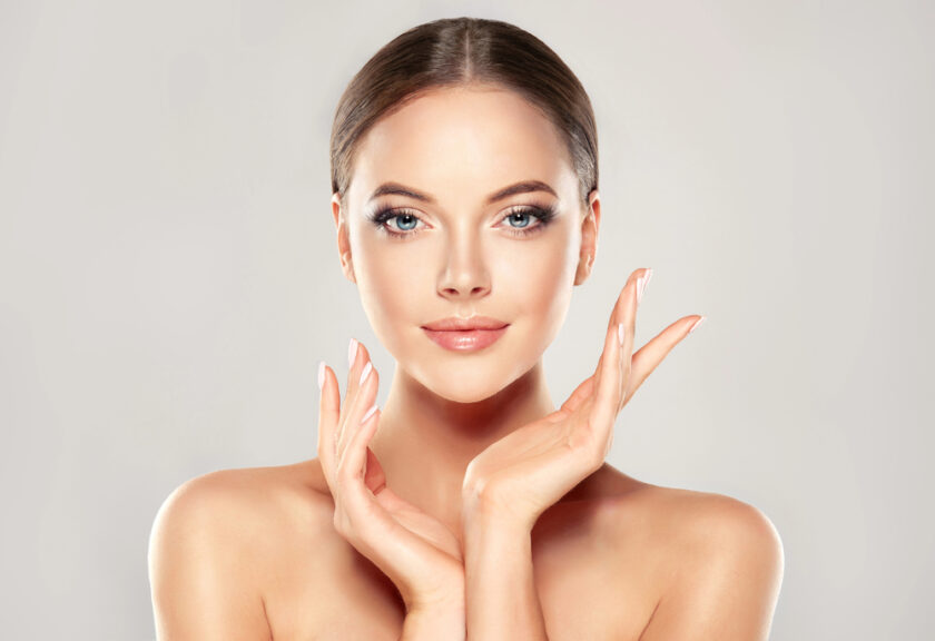 A woman with a serene expression poses with her hands gently touching her face. She has smooth skin, subtle makeup, and her hair is neatly pulled back. The background is softly blurred, adding focus to her features.
