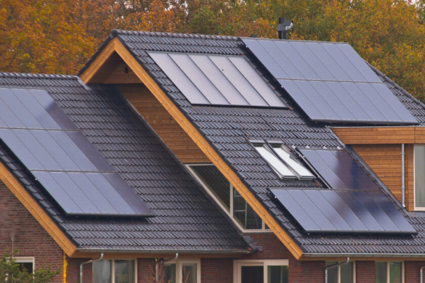 A modern house with multiple solar panels installed on its dark tiled roof. The building features wooden accents and large windows, surrounded by trees with autumn foliage in the background.