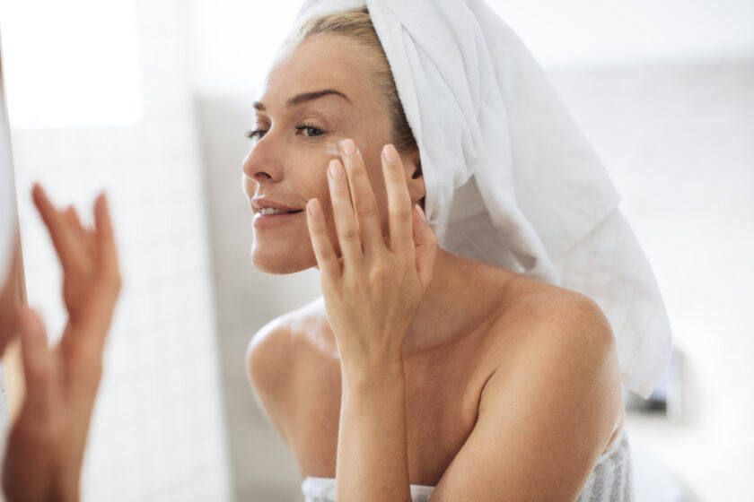 Pretty Caucasian woman looking her face in bathroom mirror and applying cosmetic.
