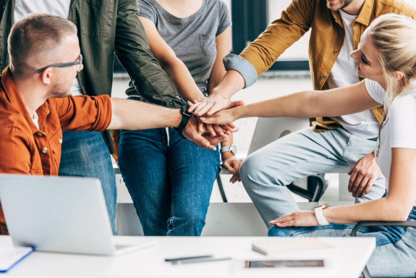 A group of diverse people stands in a circle with their hands stacked in the center, symbolizing teamwork and unity. A laptop and notebook are visible on the table nearby. Casual attire and relaxed setting suggest a collaborative environment.