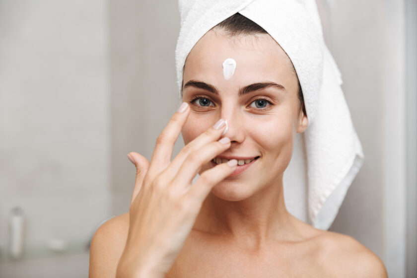 Close up of a beautiful young woman with towel on her head standing at the bathroom, applying facial cream
