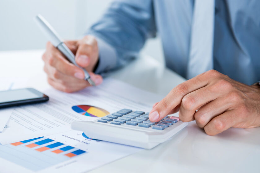 A person in a blue shirt uses a calculator and holds a pen over financial documents with charts and graphs. A smartphone is nearby on the white desk. The scene suggests financial analysis or accounting work.