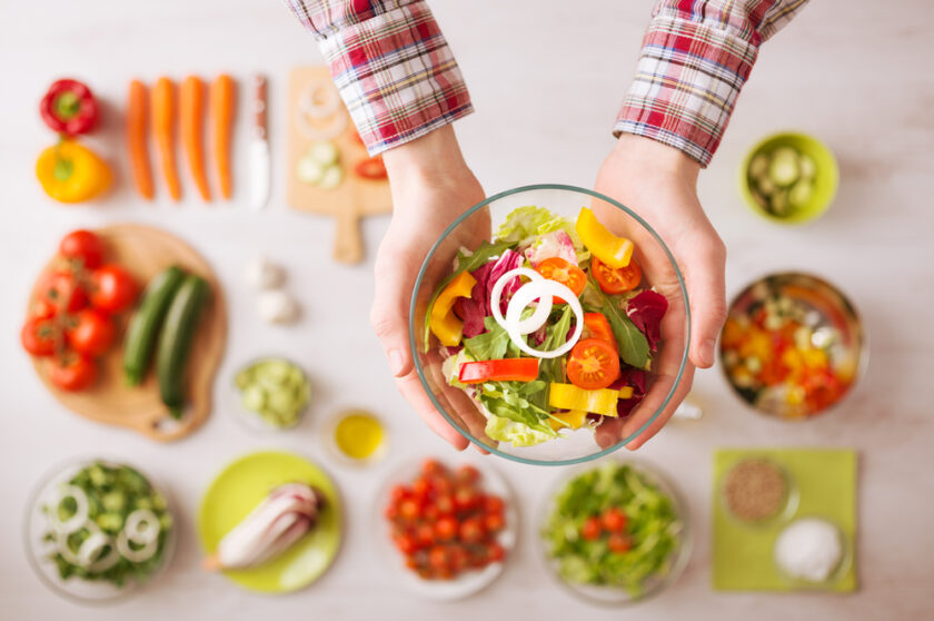 Man holding a fresh garden salad bowl with raw sliced vegetables, hands close up top view, ingredients and utensils on background