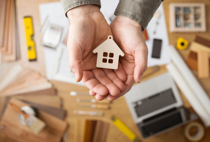Hands holding a small wooden house cutout over a table scattered with tools, a calculator, a laptop, and various wood samples, suggesting a home renovation or construction theme.