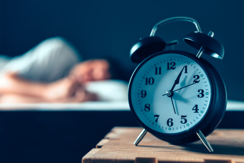Sleeping disorder or insomnia concept, selective focus of vintage clock in bedroom and out of focus male person trying to fall asleep in bed