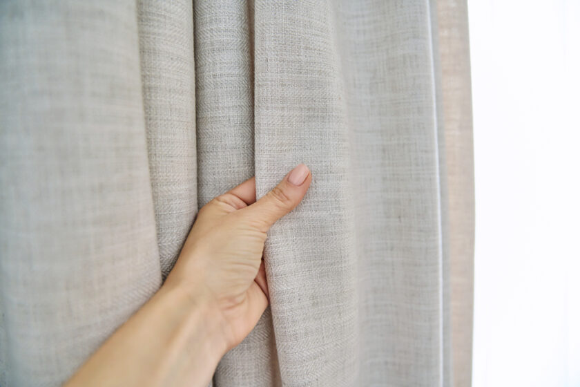 Light beige sand linen natural curtains on the window, close-up of a hand touching the curtains. Textile window decoration, fashion, style, design concept