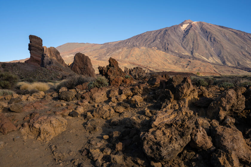 Rocky landscape with scattered volcanic rocks in the foreground and a large mountain, partially lit by sunlight, in the background under a clear blue sky. Sparse vegetation is visible among the rocks.