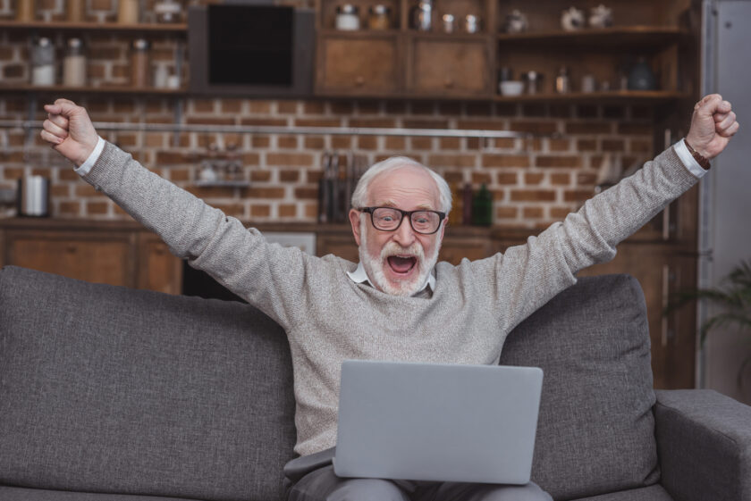 An elderly man with glasses is sitting on a gray couch, excitedly raising his arms while looking at a laptop. He appears joyful, with a brick wall and shelves in the background.