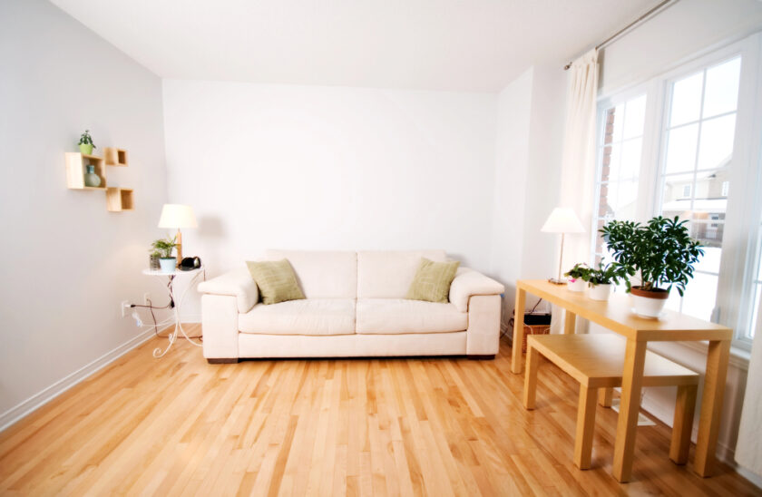 A living room with a beige couch against a white wall, flanked by a small table with plants on the right and floating shelves with decor on the left. The room has a wooden floor and is brightened by natural light from a large window.