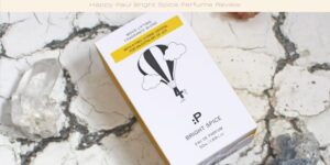 A box of Happy Paul Bright Spice Eau de Parfum is on a white marble surface. The packaging features a hot air balloon graphic and text describing the fragrance as mood-lifting and bright. Two crystal stones are nearby.