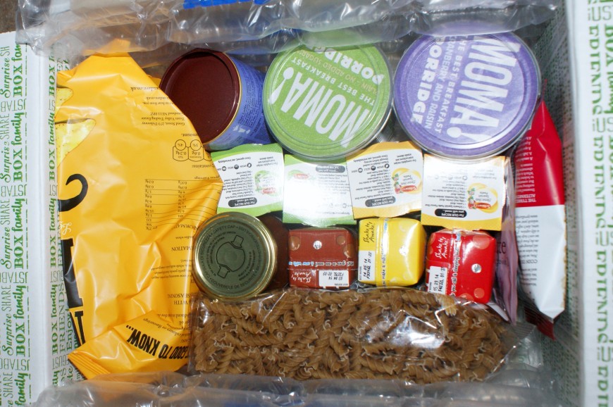 A cardboard box filled with packaged food items, including tins, small juice cartons, a bag of pasta spirals, and various sealed containers, all cushioned with inflatable packing material.