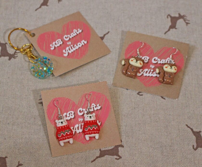 A set of handmade jewellery items on a fabric background with deer print. One keychain with a glittery orb and two pairs of earrings: one with knitted bear designs and another with brown squirrels. Displayed on cards labelled AB Crafts by Alison.