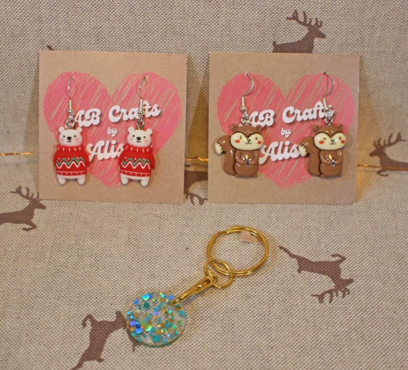 Two pairs of animal-themed earrings are displayed on cards with a pink heart design. One pair features teddy bears in red sweaters, and the other pair has squirrels. A sparkly round keychain is placed below on a fabric with deer silhouettes. Items made by AB Crafts by Alison