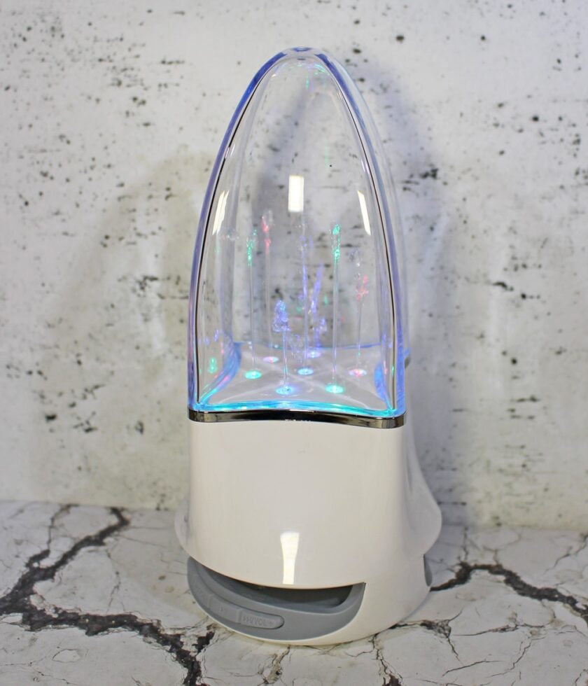 A white plastic dome-shaped speaker with a clear top emits colourful lights. The device is placed on a marbled surface against a speckled backdrop.