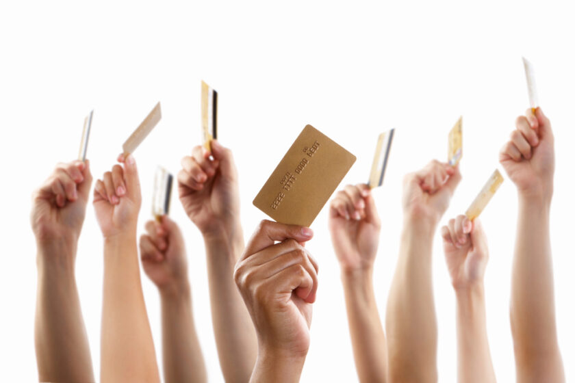 A group of diverse hands holding up credit cards against a plain white background, with only the hands and cards visible.