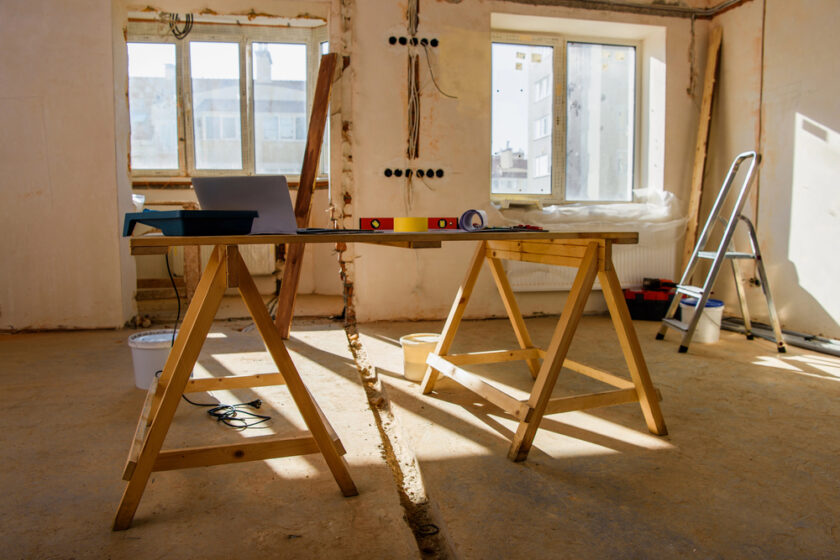 A sunlit room under renovation with a wooden sawhorse table, a laptop, and construction tools. Windows overlook neighboring buildings. A ladder and paint supplies are visible in the background.