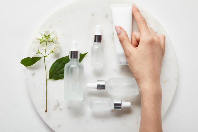 A hand reaches for a white skincare tube on a marble surface, surrounded by frosted glass dropper bottles and a sprig with white flowers and green leaves.