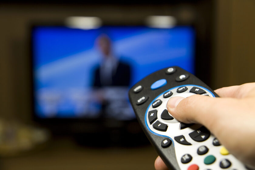 A hand holds a TV remote control, pointing it at a television screen. The TV displays a blurry image of a person, suggesting a show or broadcast in progress. The remote is black with various buttons, including a circular central section.