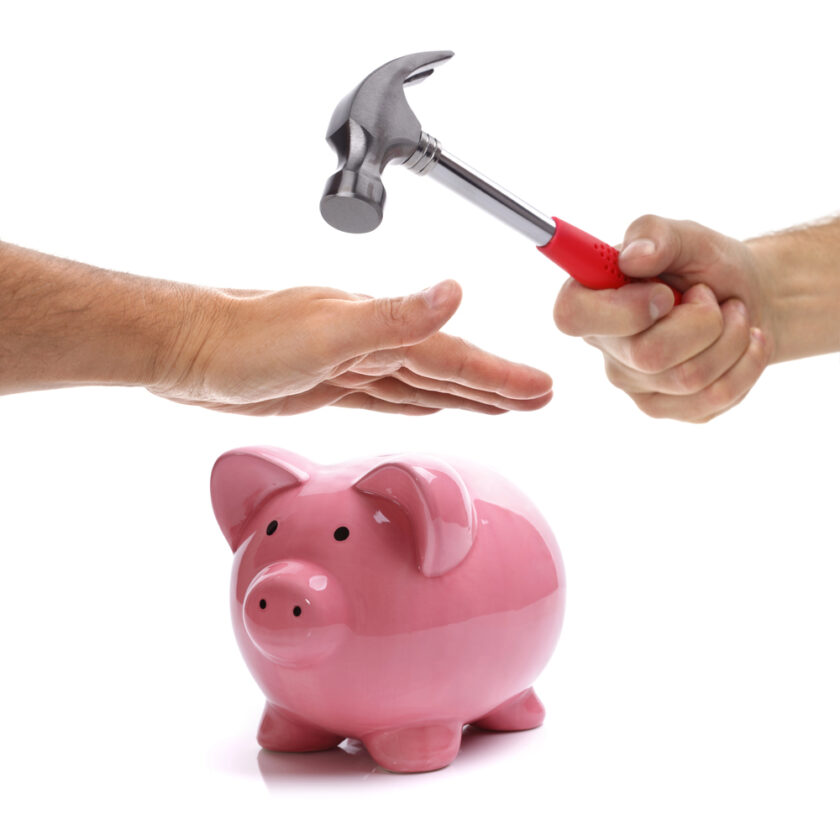 A pink piggy bank sits on a white surface. One hand holds a hammer above the piggy bank while another hand is placed protectively in front of the hammer, suggesting an attempt to prevent it from striking the piggy bank.