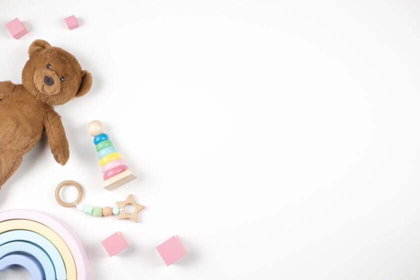 A brown teddy bear, a wooden stacking ring toy, a colorful wooden rainbow, pink blocks, and a teething ring with a bead and star shape are arranged on a white background. Ample negative space is on the right side.