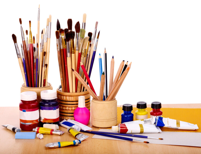 An assortment of art supplies on a wooden table, including jars filled with paintbrushes, containers holding colored pencils, paint tubes, and bottles of paint. The arrangement is bright and colorful, against a white background.