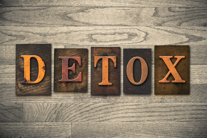The word DETOX is spelled out in large, wooden block letters on a wooden background with a textured gray pattern. Each letter is carved in a classic font style, giving a rustic and vintage feel.