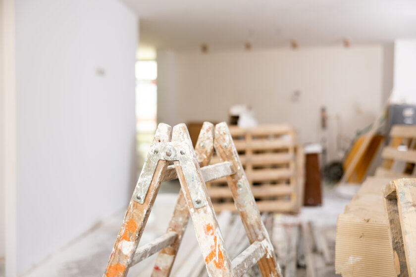 A construction ladder with paint splatters stands in a room under renovation. The background shows scattered construction materials and a partially painted wall.