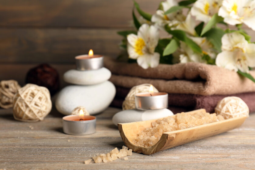 A relaxing spa scene with lit candles, stacked white stones, a bamboo tray of bath salts, woven balls, and folded towels. White flowers are in the background on a wooden surface.