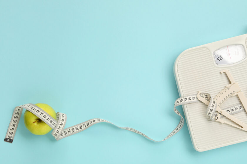 A white analog bathroom scale rests on a light blue background, accompanied by a measuring tape and a green apple. The measuring tape is wrapped around the apple and partially extends onto the scale.