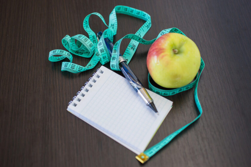 A notebook, pen, green apple, and blue measuring tape are placed on a dark wooden surface. The tape is loosely coiled around the apple, suggesting themes of health, diet, or fitness planning.