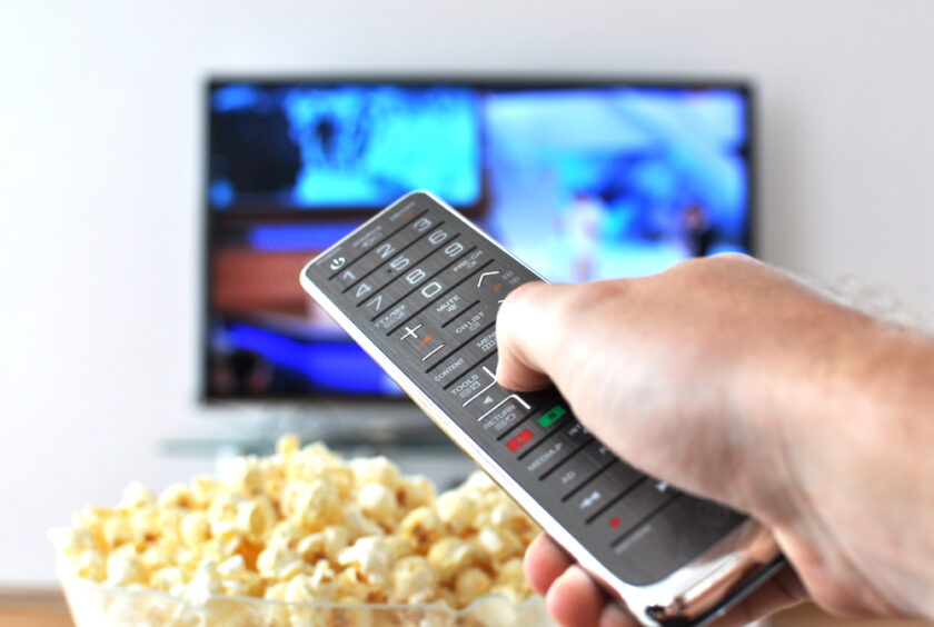 A hand holding a remote control is pointed towards a television in the background. In the foreground, theres a bowl of popcorn. The screen displays a blurred image.