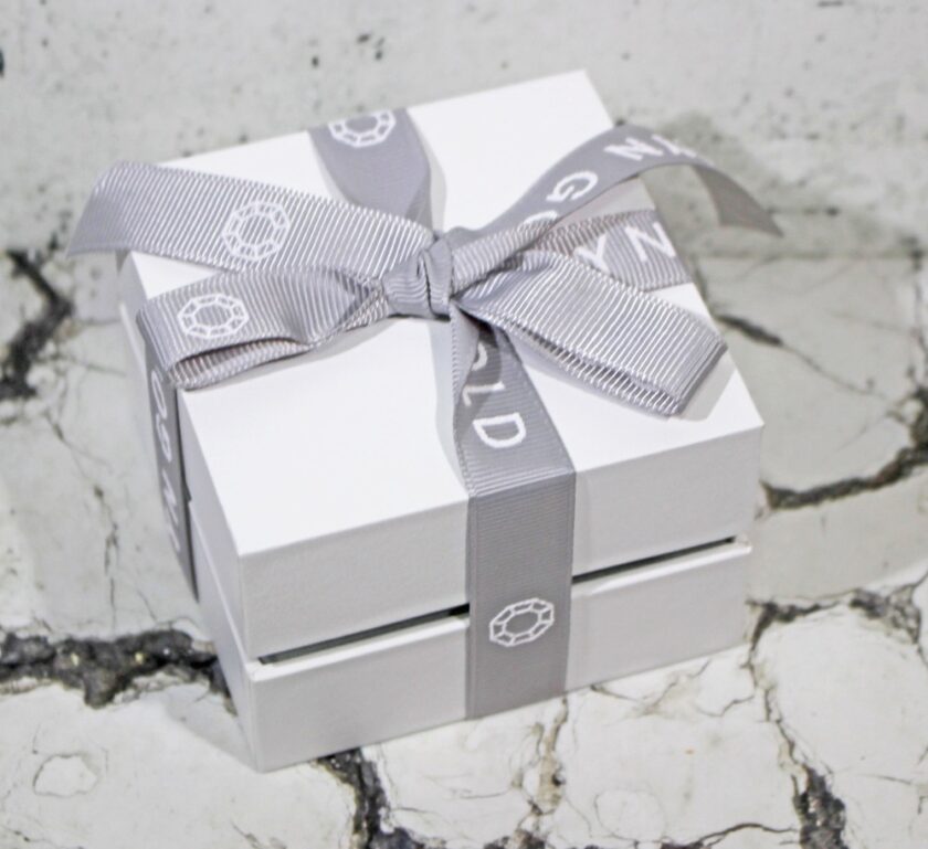 A small white gift box adorned with a light gray ribbon sits on a cracked, textured marble surface. The ribbon features patterned designs and loops elegantly into a bow on top.
