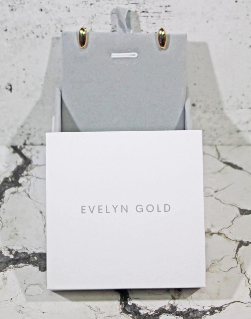 A pair of gold earrings is displayed in an open gray jewelry box on a marble surface. The box features the text EVELYN GOLD in elegant font on the front.