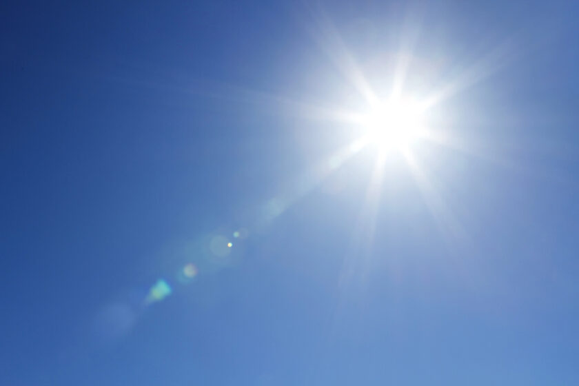 Bright sun shining in a clear blue sky, with visible lens flare and radiating light beams.