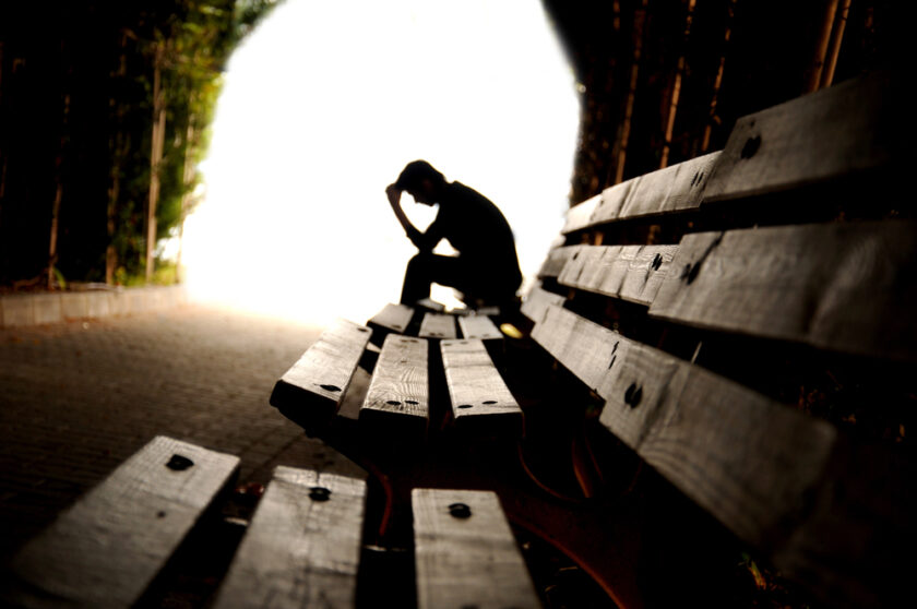 Silhouette of a person sitting on a bench in a tunnel, resting their head on one hand in a thoughtful or contemplative pose. The background is brightly lit, creating a stark contrast with the dark tunnel interior.