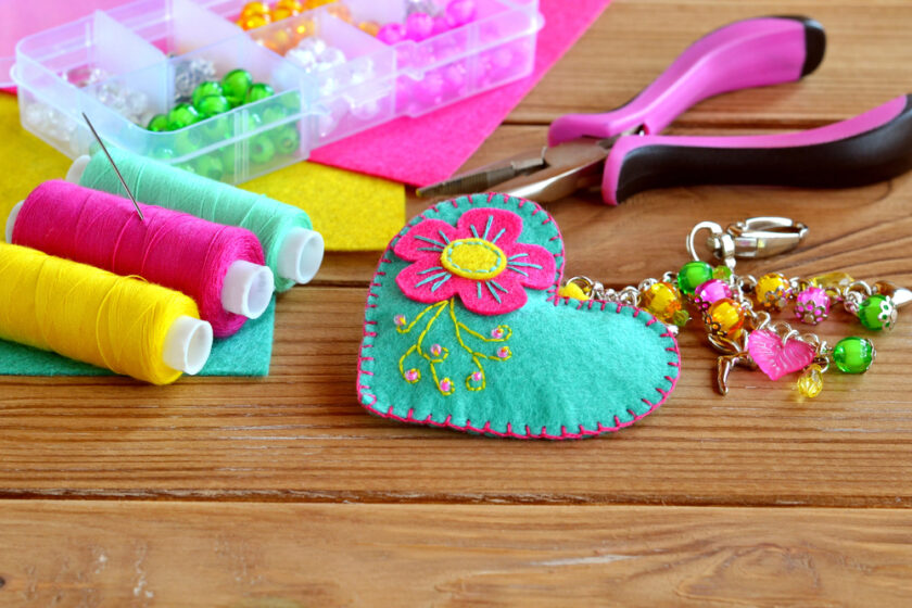 Colorful sewing supplies on a wooden table, including pink, yellow, and green thread spools, a pink heart-shaped felt ornament with floral embroidery, large pliers, and a box of beads. A keychain with beads and charms is also visible.