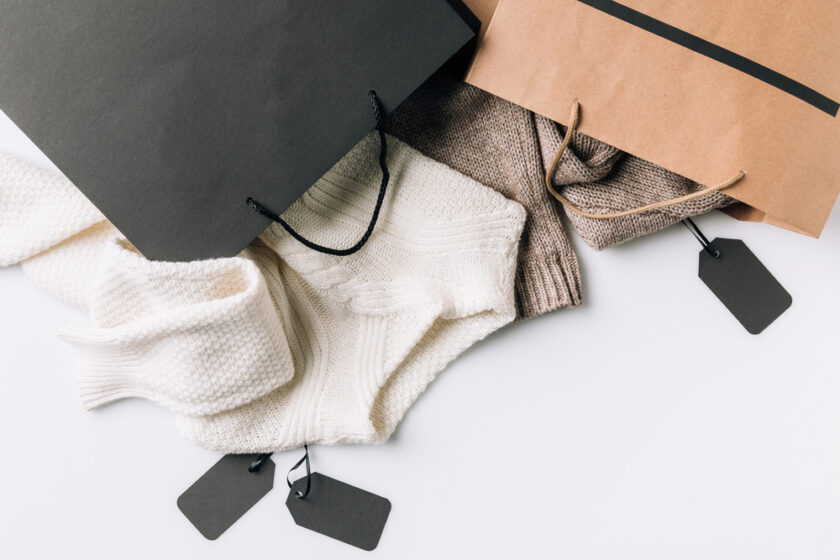 Shopping bags with cozy sweaters in white and beige partially visible. Two blank black price tags are attached to the bags, which are positioned on a white surface.