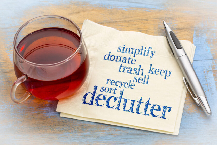 A glass cup filled with tea sits on a wooden surface. Next to it is a pen and a napkin with the words simplify, donate, trash, keep, sell, recycle, sort, declutter printed in blue.