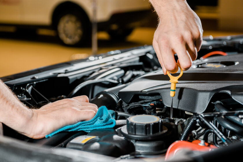 Close-up of a person checking a cars oil level with a dipstick under the hood. One hand holds the dipstick, while the other hand, resting on a blue cloth, supports the process. Car engine components are visible in the background.