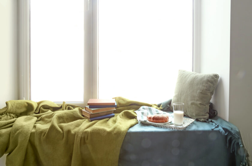 A cozy window seat with a green blanket and cushion, stacked books, a plate with a pastry, and a glass of milk. Soft light streams in through the window, creating a tranquil atmosphere.