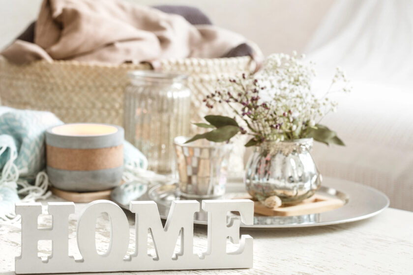 A cozy setting with a candle, a silver vase with white flowers, and decorative silver items on a table. A basket with a blanket is in the background, and a HOME sign is in the foreground, creating a warm atmosphere.