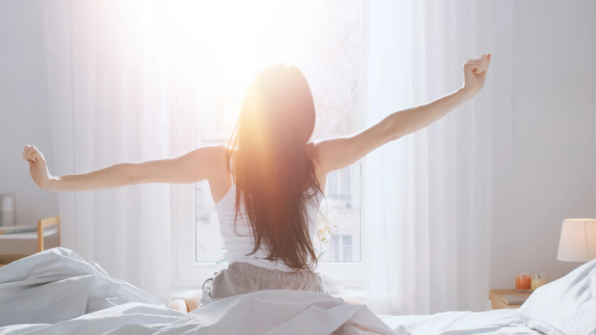 A person with long hair stretches while sitting on a bed, facing a bright window. The room has white curtains and sunlight streams in, creating a warm and serene atmosphere. A bedside lamp is visible to the right.