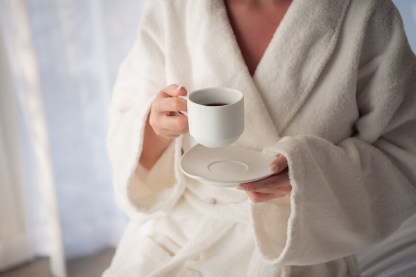 Attractive young girl in bathrobe holding a cup, looking at window and smiling while sitting on a bed near the big window at home.