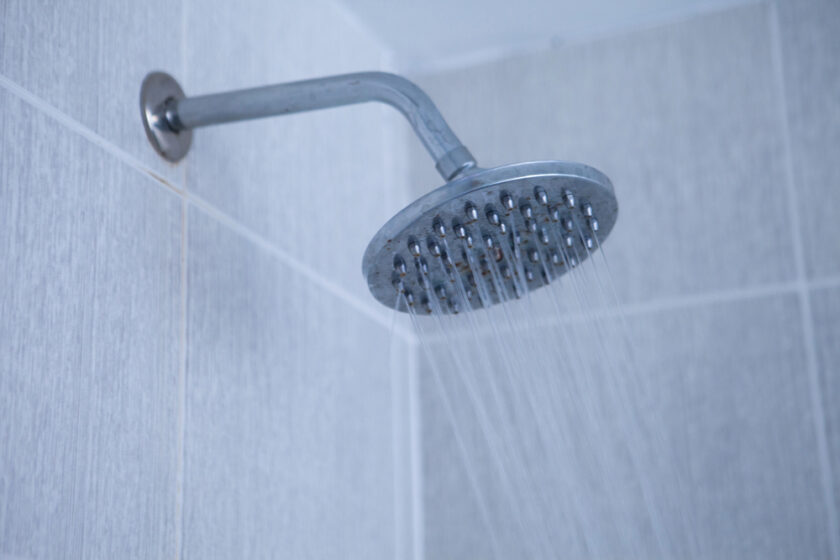 A wall-mounted showerhead sprays water in a tiled bathroom, showing droplets streaming down from its circular surface. The tiles are light gray, creating a simple and clean shower space.