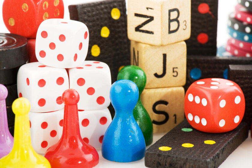 A collection of colorful board game pieces, including red and white dice, wooden letter tiles, dominoes, and multi-colored game pawns. Stacks of poker chips are also visible in the background, creating a vibrant assortment.
