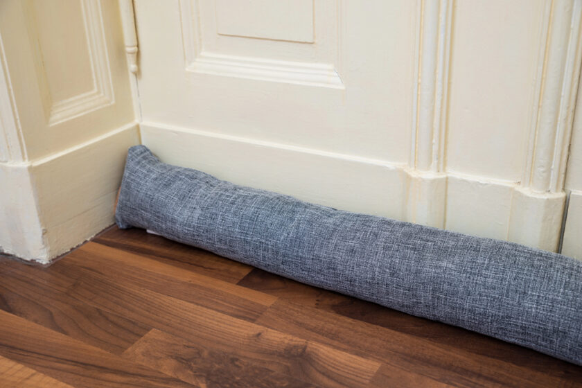 A gray fabric door draft stopper placed at the bottom of a white wooden door on a brown hardwood floor.
